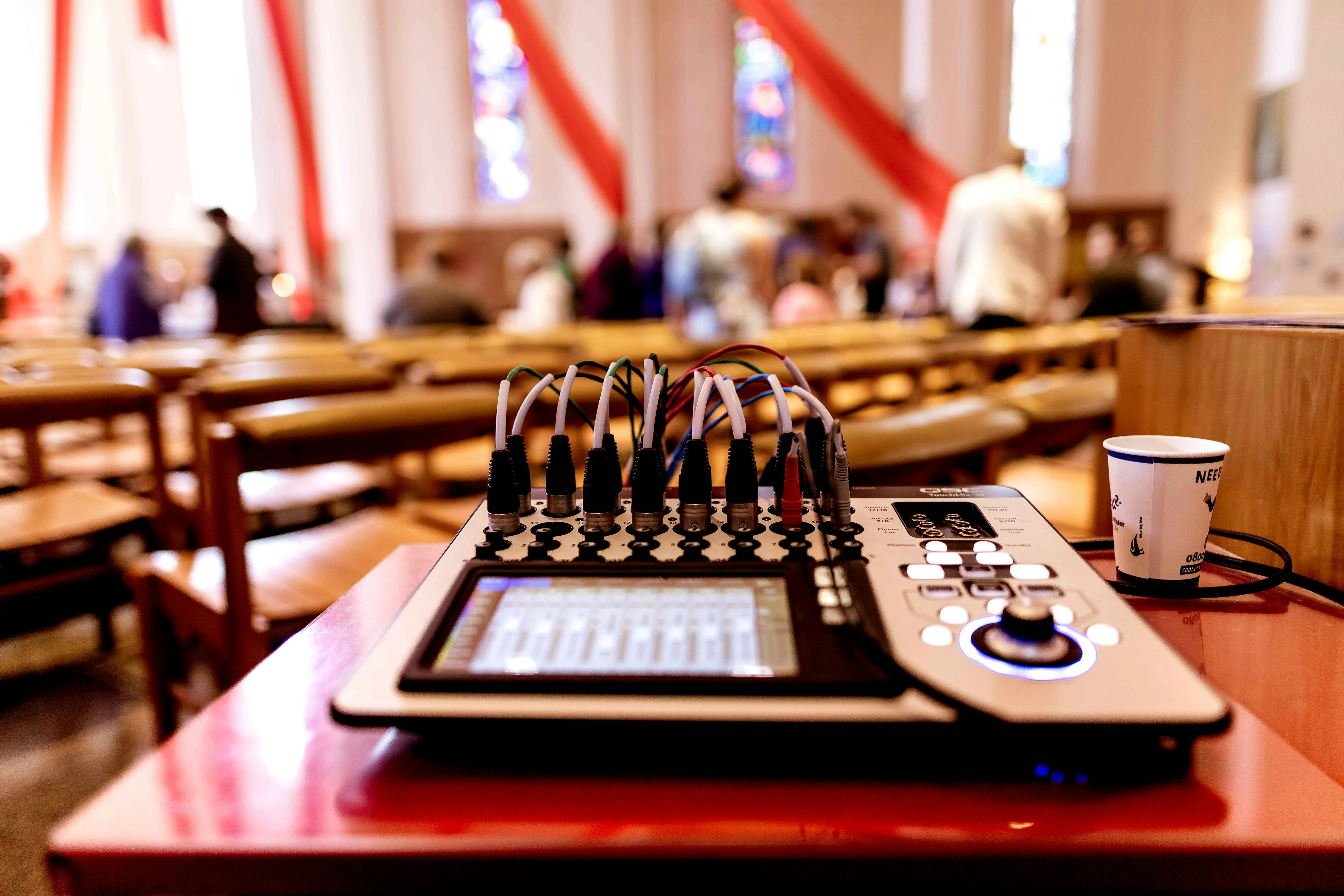 A compact digital mixer with touchscreen deployed on a church table during a live service, congregation visible in the background
